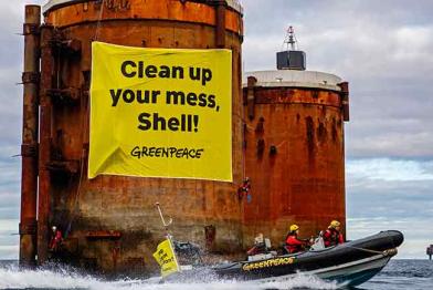 Greenpeace-Aktivisten und Aktivistinnen protestieren im Schlauchboot vor der Brent Bravo und Brent Alpha. An einem Betonsockel hängt ein Banner "Clean up your mess, Shell!"
