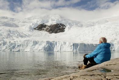 Ein See, dahinter mit Eis und Schnee überzogene Berge. Am See sitzt, mit dem Rücken zum Betrachter, der Forscher Claude Lorius