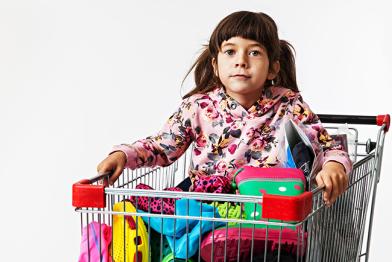 Ein kleines Mädchen sitzt in einem Einkaufswagen, der mit Klamotten gefüllt ist. 