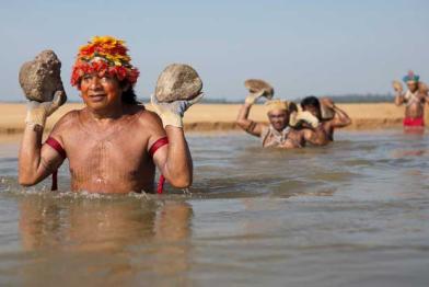 Vier Männer des Munduruk-Volkes waten in den Tapajós-Fluss hinein. Sie tragen bunte, zeremonielle Kopfbedeckungen und je zwei Steine, die sie auf den Handflächen über Wasser halten.