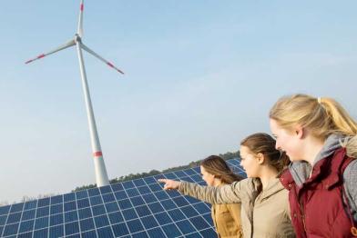 Drei junge Frauen stehen vor einem Solarpanel, dahinter dreht sich ein Windrad. Eine der Frauen zeigt auf die Anlagen.