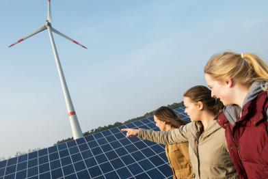Drei junge Frauen stehen vor einem Solarpanel, dahinter dreht sich ein Windrad. Eine der Frauen zeigt auf die Anlagen.