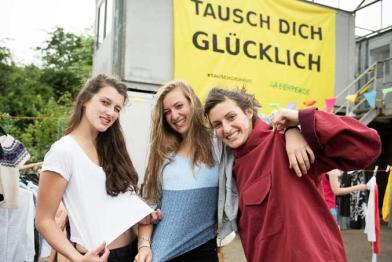 Drei Mädchen präsentieren auf der Kleidertauschparty in Hannover ihre "neuen" Stücke. Über ihnen hängt ein Banner mit der Aufschrift "Tausch dich glücklich"