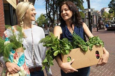 Zwei lachende junge Frauen gehen eine Straße entlang. Sie halten Pappkartons, aus denen Oben das Grün von Gemüse herausschaut.