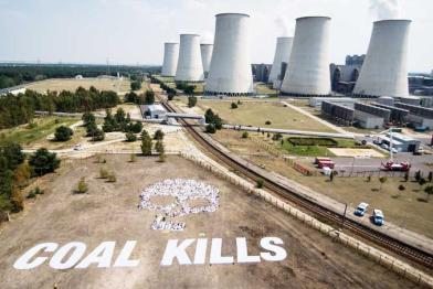 Die rauchenden Schlote des Kohlekraftwerks Jänschwalde; davor auf einer Sandfläche haben sich weiß gekleidete Aktivisten zu einem Totenkopf formiert. Darunter ist ein weißer Totoenkopf zu sehen.
