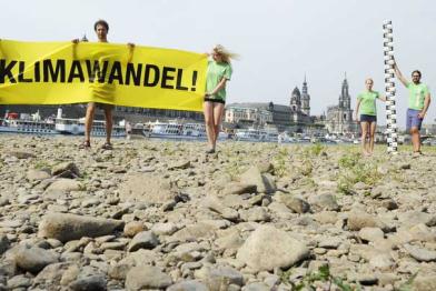 Greenpeace-Aktivisten stehen in der ausgetrockneten Elbe bei Dresden und halten eine große Pegelmesslatte und ein Banner mit der Aufschrift "Klimawandel!" hoch.
