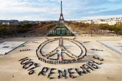 Im Pariser Park des Champ-de-Mars formen Hunderte Menschen das Eiffelturm-Friedenssymbol und darunter den Schriftzug 100% Renewable. Im Hintergrung ist der Eiffelturm zu sehen