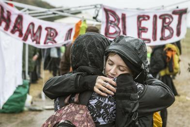 Zwei Frauen umarmen sich im Regen vor einem Banner "Hambi bleibt"