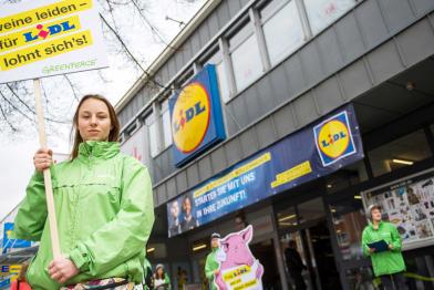 Greenpeace Ehrenamtliche protestieren vor Lidl in Hamburg gegen Billigfleisch. Eine Aktivistin trägt ein Schild mit der Aufschrift „Schweine leiden - für Lidl lohnt sich's!“. Im Hintergrund ein Schwein aus Pappe.