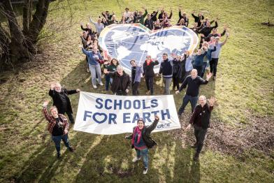 Schüler:innen und Lehrer:innen halten auf einer Wiese gemeinsam mit dem Greenpeace Schools for Earth Team das Schools for Earth Herz und ein Text Banner in die Luft und winken in die Kamera 
