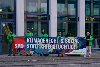 Am Eingang zum Konferenzgebäude in Berlin halten Aktivist:innen ein 5 Meter breites Banner in Camouflagemuster mit der Aufschrift: „ Klimagerecht und sozial statt kriegstüchtig!“