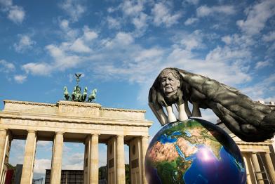 Weltkugel mit Merkel-Konterfei unter Kohlewolken vorm Brandenburger Tor in Berlin