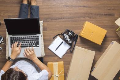 Mensch mit Laptop sitzt auf dem Boden inmitten von Paketen