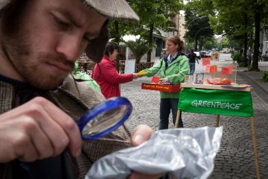 Greenpeace-Aktivisten protestieren im und vor dem KaDeWe (Kaufhaus des Westens) gegen den Verkauf von US-Lebensmitteln, die gentechnisch veränderte Zutaten enthalten. Gen-Detektiv schaut mit einer Lupe auf die kleingedruckte Zutatenliste eines Produkts.