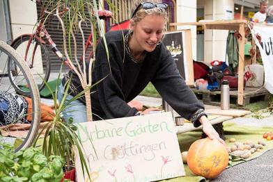 Park(ing) Day: eine Frau breitet einen Kürbis, Kartoffeln und Tomaten auf einer Decke aus, wo sonst Autos parken.