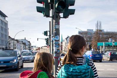 Zwei Schulkinder stehen an einer Straßenkreuzung mit viel Verkehr.