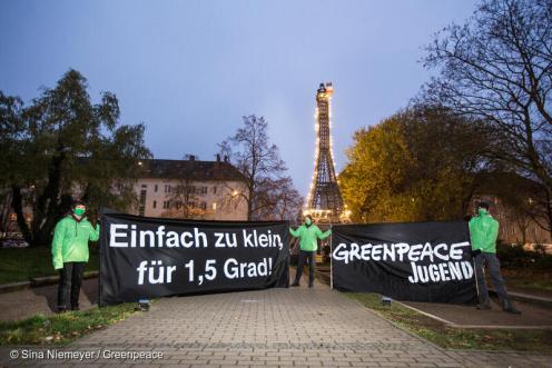 Zu sehen sind drei Jugendliche die zwei Banner halten: Einfach zu klein für 1,5 Grad und Greenpeace Jugend. Im Hintergrund steht ein brennendes Modell des Eifelturms
