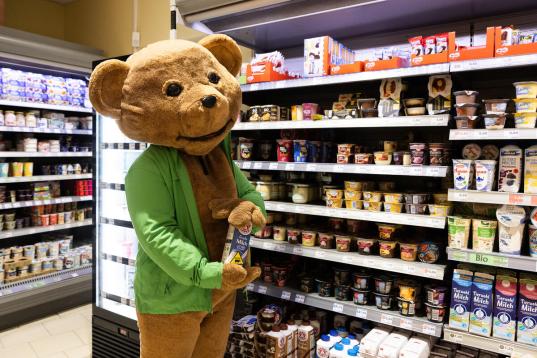 Mensch in Bärenmarkekostüm steht im Supermarkt vor einem Kühlregal mit Milchprodukten und hält eine mit Aufkleber versehene Bärenmarke-Milchpackung "Achtung Tierleid"