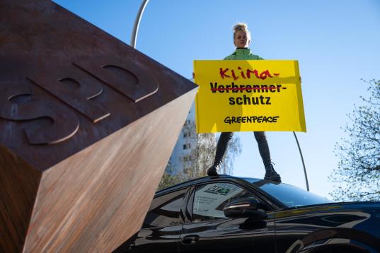 Aktivistin steht auf einem Auto mit einem Banner: Beim Wort Verbrenner-schutz ist das Wort "Verbrenner" durchgestrichen und stattdessen "Klima" reingesetzt
