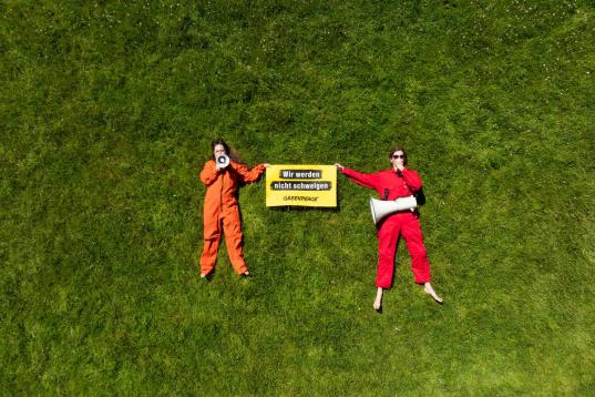 Zwei Menschen im Gras mit Greenpeace-Protestplakat "Wir werden nicht schweigen" und Megafon