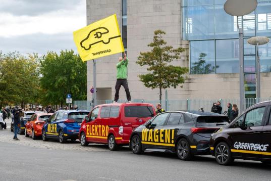 Ein Aktivist steht mit einer Fahne mit stilisiertem E-Auto auf einem Autodach. Drei Autos formen zusammen die Aufschrift “Mehr Elektro Wagen”.