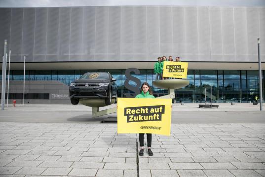 Eine überdimensionierte Waage: auf der einen Seite der schwerer wiegende VW, auf der anderen Menschen, die ein Banner halten mit der Aufschrift 2Recht auf Zukunft