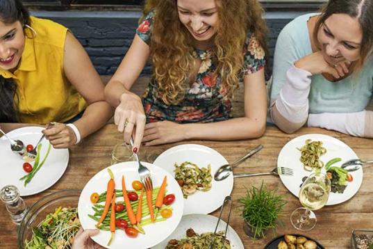 Drei junge Frauen sitzen an einem mit Getränken und vegetarischen Speisen gedeckten Tisch.