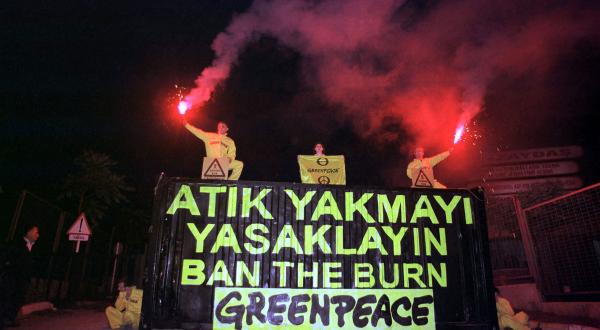 Greenpeace-Aktivist:innen nachts vor dem Eingang der Verbrennungsanlage Izaydas in der Westtürkei. Sie halten brennende Fackeln über einem schwarzen Container mit der Aufschrift "Ban the burn". 