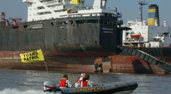 Greenpeace-Aktivist:innen in einem Schlauchboot rasen an zwei illegalen Schiffen, darunter der 1980 gebauten 'Genova Bridge' aus Großbritannien vorbei, die potenziell sie giftige Substanzen an Bord haben.