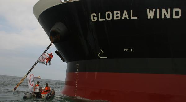 Greenpeace-Person, angekettet an die Ankerkette des Massengutfrachters Global Wind, mit einem Banner mit der Aufschrift (durchgestrichen): "GMO" mit Greenpeace-Schlauchboot, Paranagua, Brasilien. 