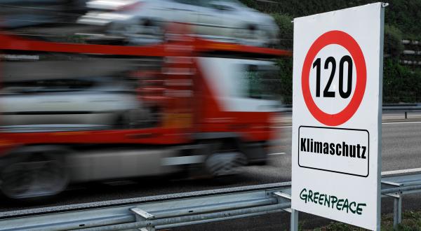 An einer Straße steht ein Schild "Tempo 120. Klimaschutz. Greenpeace".  Ein LKW fährt vorbei.