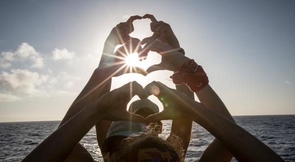 Mitglieder der Greenpeace Esperanza-Crew formen Herzen mit ihren Händen, im Hintergrund ist das Meer zu sehen. 