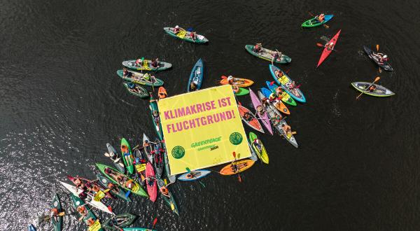 Kajaks, Kanus und Schwimmer:innen im Wasser, auf einem schwimmenden Banner steht: Klimakrise ist Fluchtgrund!