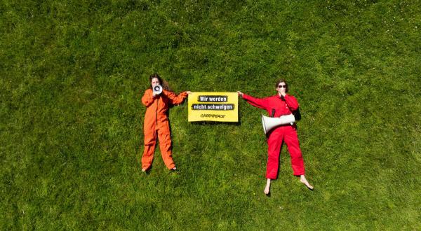Zwei Menschen im Gras mit Greenpeace-Protestplakat "Wir werden nicht schweigen" und Megafon