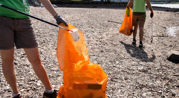 Zwei Aktive sammeln mit Greifzangen Müll am Flussufer, im Hintergrund Häuser einer Stadt