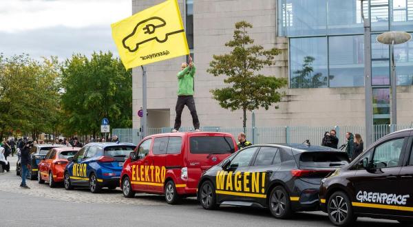 Ein Aktivist steht mit einer Fahne mit stilisiertem E-Auto auf einem Autodach. Drei Autos formen zusammen die Aufschrift “Mehr Elektro Wagen”.