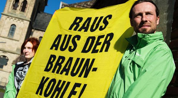 Ein Mann in grüner Greenpeace-Jacke hält gemeinsam mit einer Frau ein gelbes Plakat hoch, auf dem steht: " Raus aus der Braunkohle!"