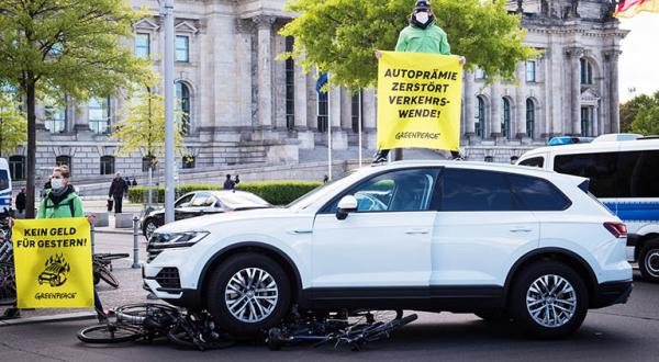 Aktivist*innen stehen auf dem Autodach und halten ein Banner "Autoprämie zerstört Verkehrswende", vor dem Auto liegen überfahrene bereits vor der Aktion schrottreife Räder.