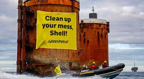 Greenpeace-Aktivisten und Aktivistinnen protestieren im Schlauchboot vor der Brent Bravo und Brent Alpha. An einem Betonsockel hängt ein Banner "Clean up your mess, Shell!"