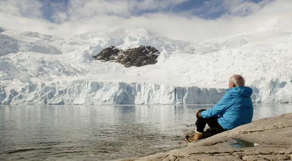 Ein See, dahinter mit Eis und Schnee überzogene Berge. Am See sitzt, mit dem Rücken zum Betrachter, der Forscher Claude Lorius