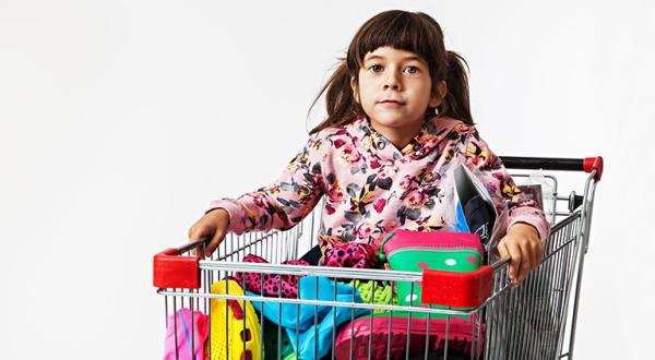 Ein kleines Mädchen sitzt in einem Einkaufswagen, der mit Klamotten gefüllt ist. 