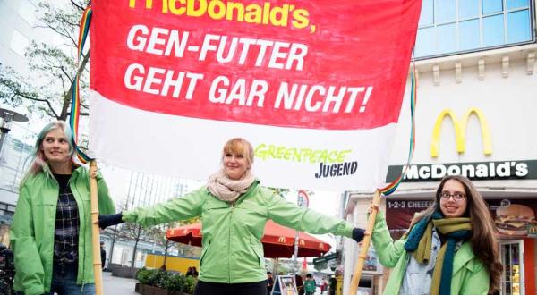 Drei Aktivistinnen der Greenpeace-Jugend stehen vor einer McDonald's-Filiale. Sie halten ein rotes Transparent mit der Aufschrift: "Gen-Futter geht gar nicht"