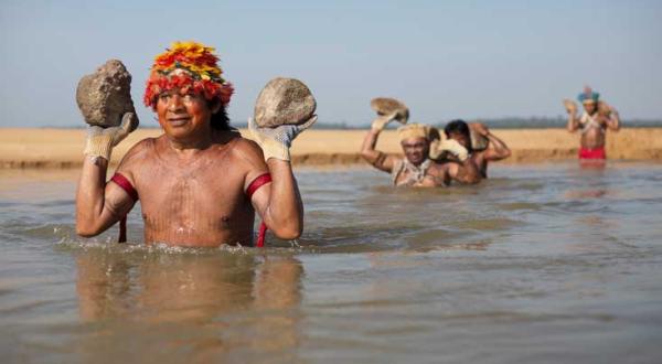Vier Männer des Munduruk-Volkes waten in den Tapajós-Fluss hinein. Sie tragen bunte, zeremonielle Kopfbedeckungen und je zwei Steine, die sie auf den Handflächen über Wasser halten.