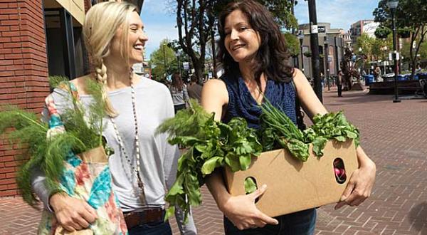 Zwei lachende junge Frauen gehen eine Straße entlang. Sie halten Pappkartons, aus denen Oben das Grün von Gemüse herausschaut.