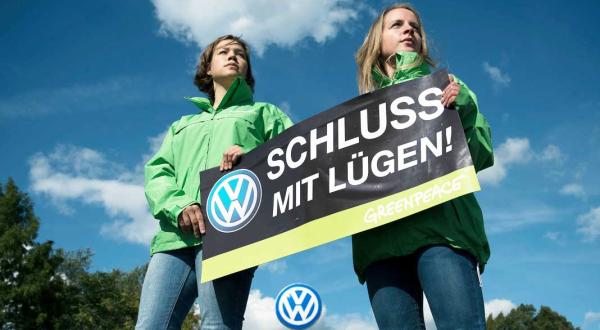 Zwei Greenpeace-Aktivistinnen in grünen Jacken stehen vor einem VW-Logo. Die beiden halten ein Banner mit der Aufschrift "Schluss mit Lügen!"; das Schild zeigt ebenfalls ein VW-Logo