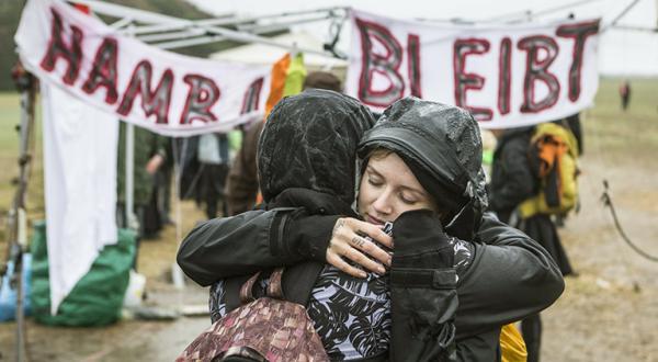 Zwei Frauen umarmen sich im Regen vor einem Banner "Hambi bleibt"