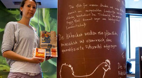 Frau steht neben McDonald's-Werbetafel. Darauf steht: „Die Hähnchen werden ohne genmodifizierte Futtermittel aufgezogen.“