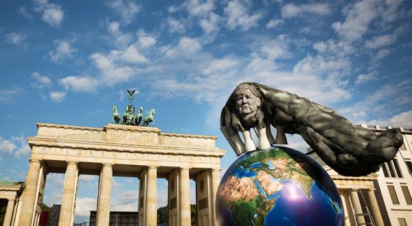 Weltkugel mit Merkel-Konterfei unter Kohlewolken vorm Brandenburger Tor in Berlin
