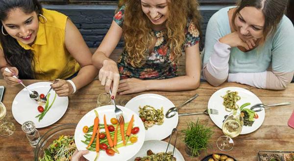 Drei junge Frauen sitzen an einem mit Getränken und vegetarischen Speisen gedeckten Tisch.