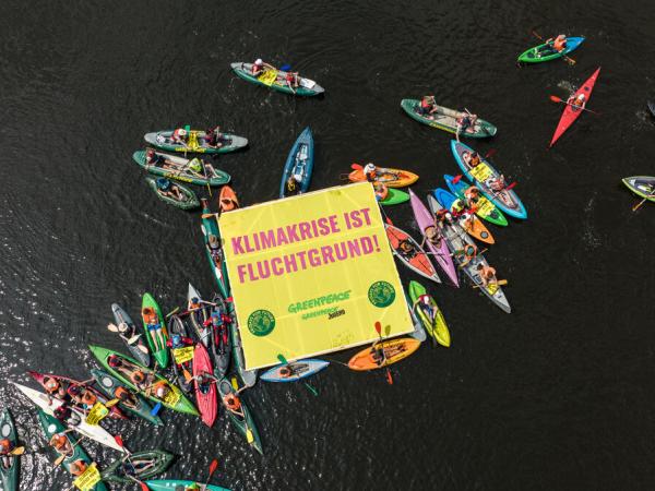 Kajaks, Kanus und Schwimmer:innen im Wasser, auf einem schwimmenden Banner steht: Klimakrise ist Fluchtgrund!
